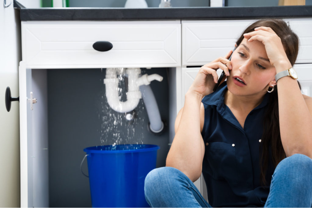 Woman sitting on the floor in front of a leaking sink making a phone call